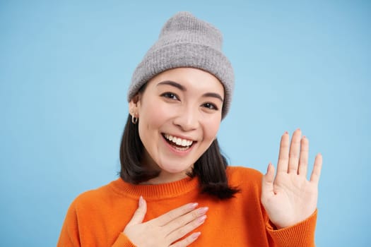 Close up of cute korean girl in beanie, raises one hand and introduces herself, greets you, smiles and looks friendly, blue background.