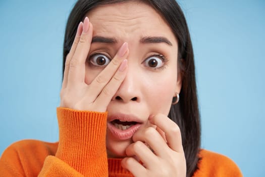 Portrait of shocked korean girl, gasping and looking worried, frowning, looks scared and anxious, stands over blue background.