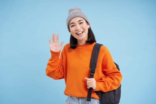 Friendly girl says hello, holds backpack and waves hand, greets you and laughs, stands over blue background.