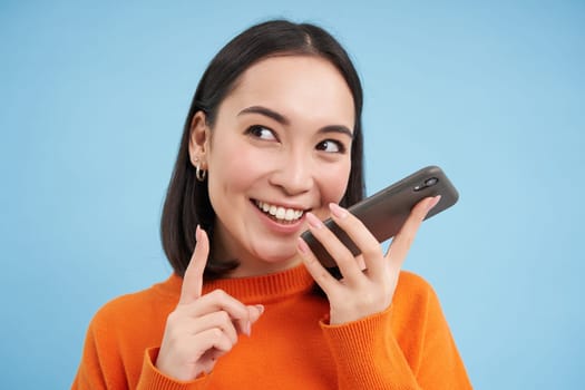 Portrait of korean woman records voice message on smartphone app, talks on speaker, translates her speech on mobile app, blue background.