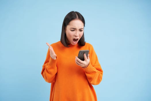 Angry korean girl swearing and shouting at mobile phone, looks outraged, furious while reading news on smartphone, standing over blue background.