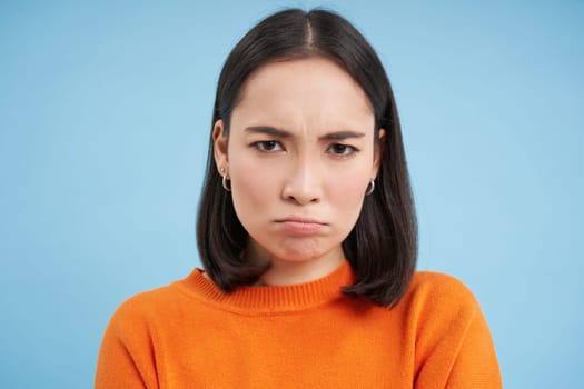 Close up of sad and gloomy japanese girl, sulking and looking upset, stands over blue background.