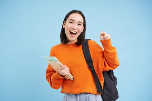 Cheerful asian student, girl with backpack and notebooks, going to college, looks happy, blue background.