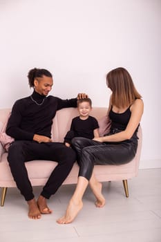 Happy multiracial young family with little boy, in black clothes, sits in the light living room on light pink sofa. Vertical. Copy space