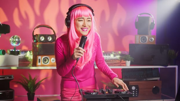 Musician having fun performing techno music at party in nightclub, using professional audio equipment. Asian performer with pink hair mixing electronic sound using mixer console. Dj concept