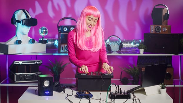 Musician with pink hair standing at dj table dancing and having fun while mixing stereo sounds with electronics. Smiling artist enjoying to perform music using professional mixer console in nightclub