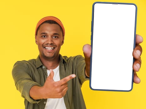 Smiling African student poses with large mobile phone featuring white blank screen, on yellow background with copy space. This image is perfect for product placement or advertising concepts. High quality photo