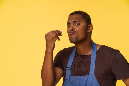 Stylish African American cook wearing blue apron sends kiss with his fingers in Italian manner, against bright yellow background with copy space for text or product placement. High quality photo