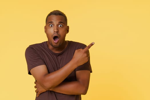 Amazed African American man is shown pointing to an empty copy space on a yellow background. This image is perfect for product placement or advertising concepts. High quality photo