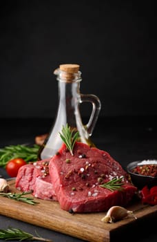 A raw piece of beef with spices, pepper, rosemary, salt, and olive oil on a wooden board, against a black background.