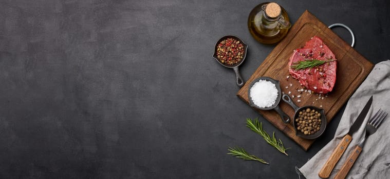 Raw piece of beef with spices pepper, rosemary sprig, salt and olive oil on a wooden board, black background. Copy space