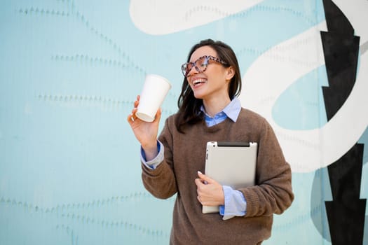 Positive business woman wearing glasses with leopard print and sweater walking down city street, holding coffee in paper cup mug and tablet computer