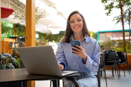 Freelance worker doing remote work using smartphone and laptop, looking at camera and smile. Female freelancer with mobile phone in outdoor cafe, restaurant on social media app online.