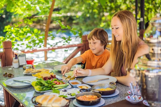 Mom and son eat Turkish breakfast. Turkish breakfast table. Pastries. Vegetables. Olives. Cheeses, fried eggs. Jams, tea in copper pot and tulip glasses. Wide composition.