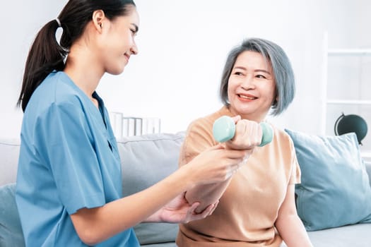 Caregiver helping contented senior woman exercise with dumbbell at home. Rehab for elder, senior medical care.