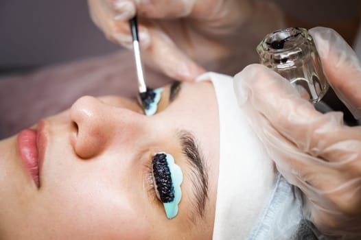 Close-up portrait of a woman on eyelash lamination procedure. The master applies tint to the eyelashes