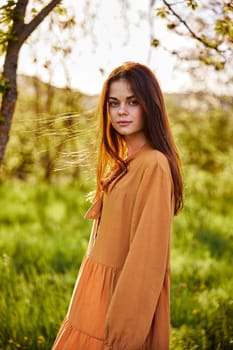 beautiful, a woman stands in a long orange dress, in the countryside, near a flowering tree, during sunset, illuminated from behind and looks sweetly into the camera. High quality photo