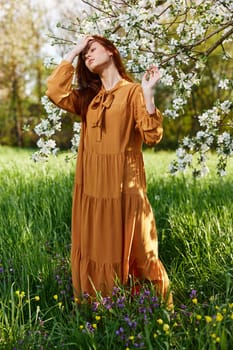 a bright vertical photo of an attractive woman in a long orange dress standing next to a flowering tree in sunny, warm weather, straightening her hair with her hand and closing her eyes. High quality photo