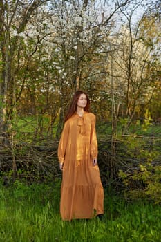 an elegant, sophisticated woman poses relaxed standing near a wicker fence at the dacha in a long orange dress. High quality photo