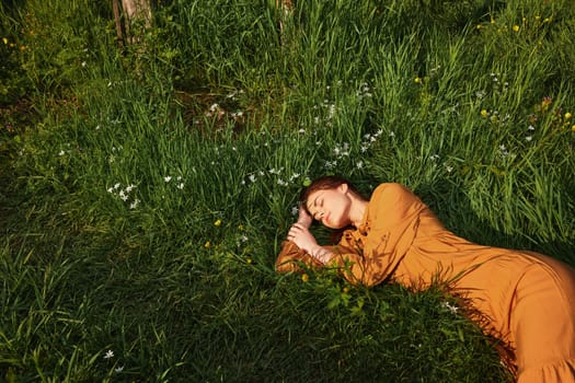 horizontal photo of a happy woman lying in the grass in a long orange dress, with her eyes closed and a pleasant smile on her face, with her hand behind her head. High quality photo