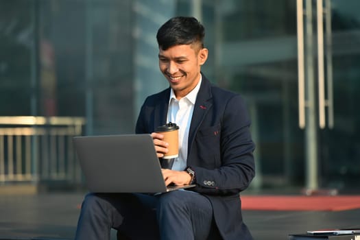 Smiling milennial man worker using laptop on stairs in front of office building. Modern lifestyle, business, technology concept.