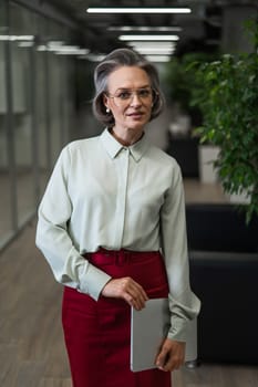 Attractive mature caucasian woman holding laptop while standing in office