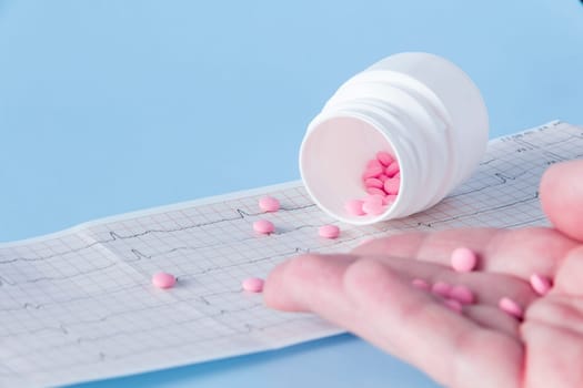 A hand pours a handful of pink pills from a white jar onto an electrocardiogram of the heart, on a blue background. The concept of a healthy lifestyle and timely medical examination