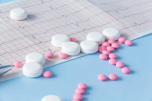 A large handful of pink and white medicines lie on the electrocardiogram of the heart, on a blue background. The concept of a healthy lifestyle and timely medical examination.