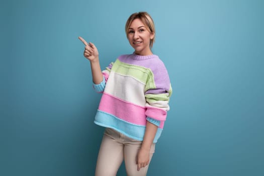 smiling blond young woman in a striped sweater points to an empty space on a blue background.