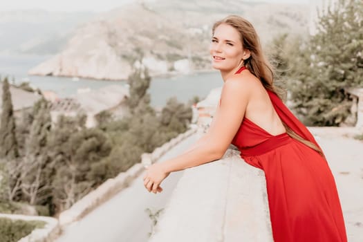 Side view a Young beautiful sensual woman in a red long dress posing on a volcanic rock high above the sea during sunset. Girl on the nature on overcast sky background. Fashion photo