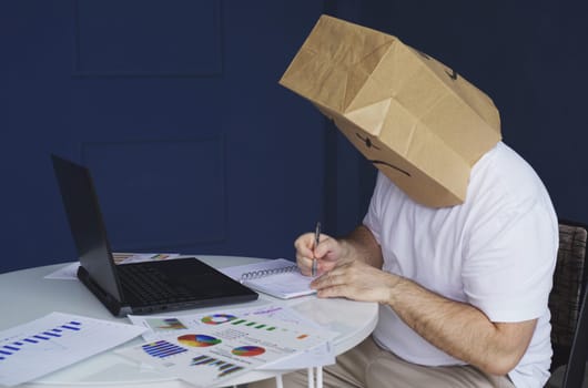 A man - a businessman in a white shirt with a paper bag on his head, with a sad smiley face drawn, works at a computer with documents, makes notes in a notebook. Emotions and gestures.
