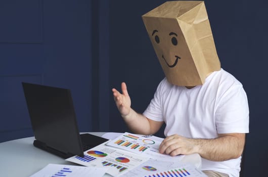 A man is a businessman in a white shirt with a paper bag on his head, with a joyful smiley face drawn, conducts a video conference or training via video link on a laptop. Emotions and gestures.