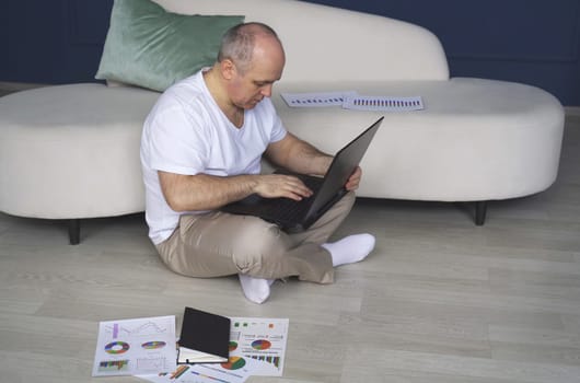 A man is sitting on the floor near the sofa, working on a laptop.