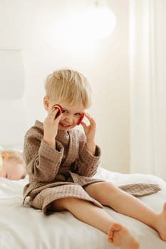 A boy in a brown bathrobe sits on the bed, plays - he closed his eyes with candied oranges. Vertical frame.