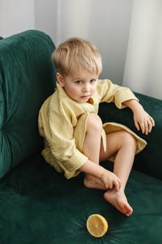 A boy in a yellow bathrobe sits on a green sofa, half an orange lies next to him.
