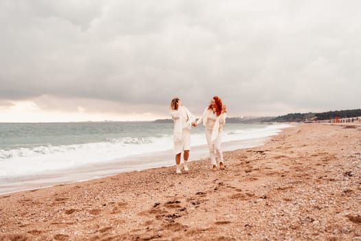 Women sea walk friendship spring. Two girlfriends, redhead and blonde, middle-aged walk along the sandy beach of the sea, dressed in white clothes. Against the backdrop of a cloudy sky and the winter sea. Weekend concept