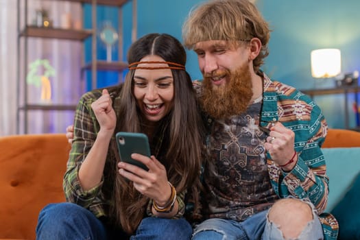 Family married hippie couple use mobile smartphone found out great big win good news celebrate lottery jackpot lucky victory, playing game, shopping online at home. Husband and wife together on sofa