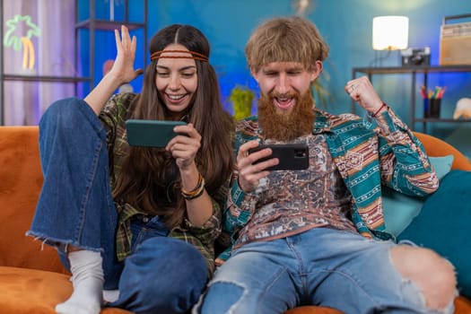 Happy family married hippie couple man woman enthusiastically playing racing or shooter video app games online on smartphone at home. Husband and wife cheating together while playing on sofa in room