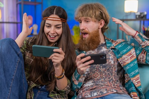 Happy family married hippie couple man woman enthusiastically playing racing or shooter video app games online on smartphone at home. Husband and wife cheating together while playing on sofa in room