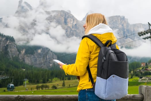 Female tourist in yellow jacket and blue backpack enjoying the view of the Dolomites Alps covered by clouds. A woman holding a mobile phone and checking the route.