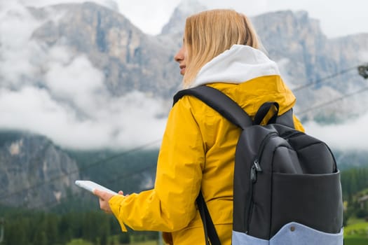 A blonde hair woman in a yellow sport jacket and blue backpack checks the route using a smartphone in Dolomites Alps