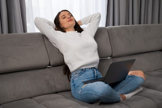 Relaxed young woman lounges on a comfortable sofa, arms crossed behind her head and holding laptop on her knees in a cozy modern living room.