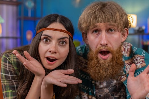Oh my God, Wow. Hippie family marriage couple man woman surprised looking at camera with big eyes, shocked by sudden victory, good win news, celebrating at home. Husband and wife together on room sofa