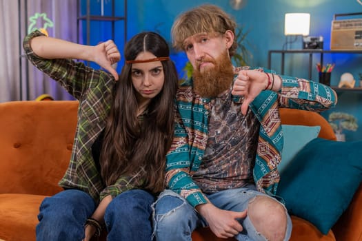 Dislike. Upset family marriage hippie couple man woman showing thumbs down sign gesture, expressing discontent, disapproval, dissatisfied bad work at home. Displeased husband and wife on sofa in room