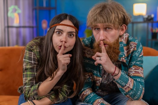 Shh be quiet please. Family marriage hippie couple man woman presses index finger to lips makes silence hush gesture sign do not tells gossip secret. Husband and wife together on sofa in living room