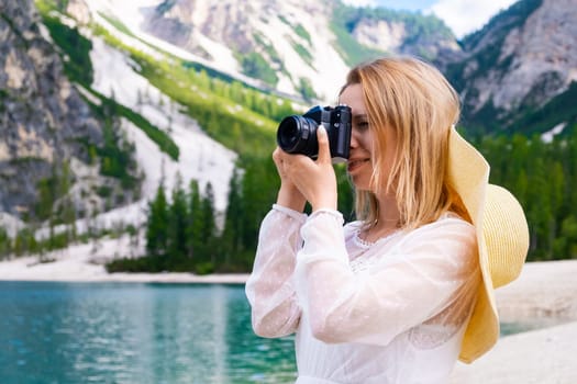 Female photographer taking photo of landscape in Dolomites Alps, Italy. Beautiful young woman in a summer hat and white dress against mountain and lake.