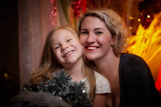 Cute mother and daughter in a room with Christmas garlands. The tradition of decorating the house and dressing up for the holidays. Gifts for family. Happy childhood and motherhood