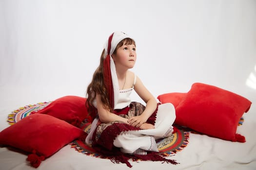 Portrait of Little girl in a stylized Tatar national costume having rest with a red pillow on a white background in the studio. Photo shoot of funny young teenager who is not a professional model
