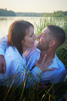 Beautiful adult couple having hugs and fun on nature in the water of a river or lake in the summer evening at sunset. A guy and a girl swim and relax outdoors in clothes in white shirts and jeans