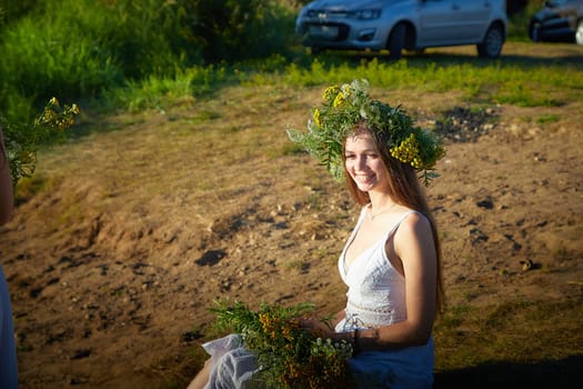 Young brunette girl in a white dress, sundress and wreath of flowers in summer on the coast of river or lake in the evening at sunset. Celebration of the Slavic pagan holiday of Ivan Kupala